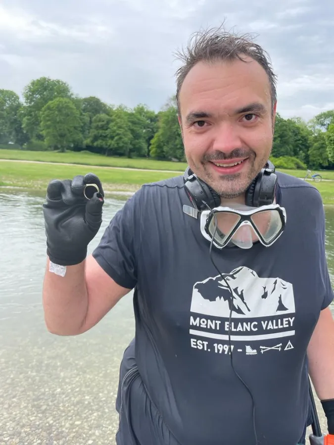 Sucher hält den geborgenen goldenen Siegelring in der behandschuhten Hand, im Hintergrund der Schwabinger Bach im Englischen Garten