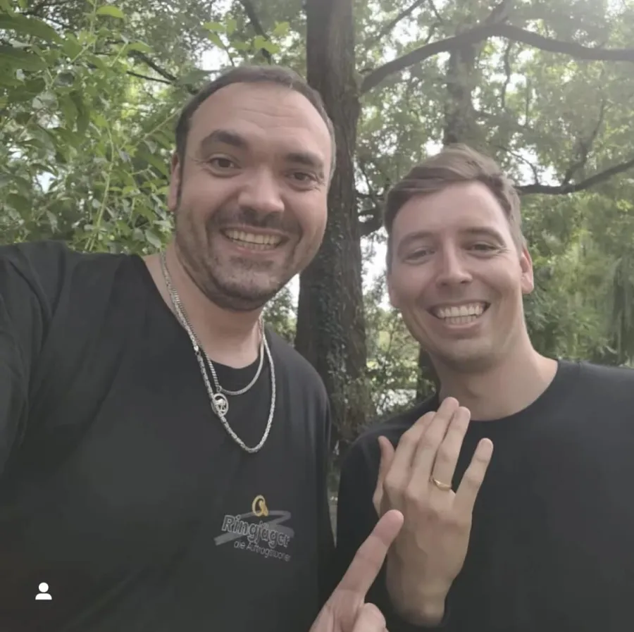Zwei Männer im Englischen Garten, einer trägt ein Ringjäger-T-Shirt und zeigt auf den wiedergefundenen Ehering am Finger des anderen