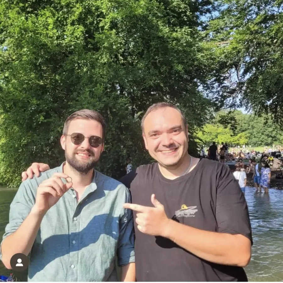 Zwei Männer am Eisbach im Englischen Garten an einem belebten Sommertag, einer hält den geborgenen Ring in der Hand