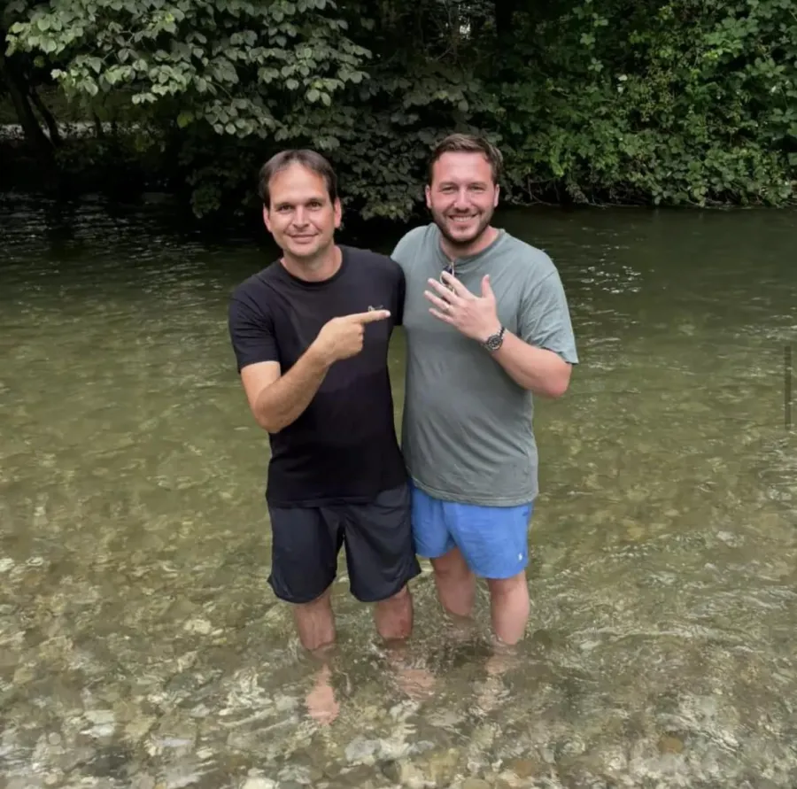 Zwei Männer stehen im flachen, klaren Wasser des Schwabinger Bachs, einer zeigt den wiedergefundenen Ehering am Finger