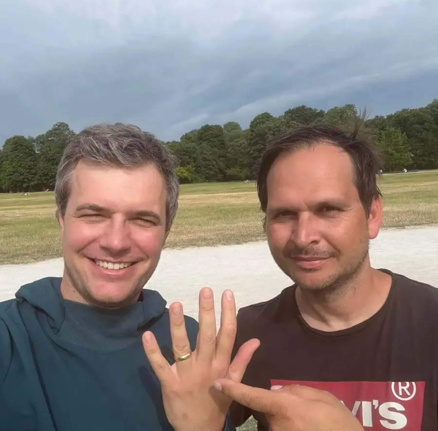 Zwei lächelnde Männer im Englischen Garten, einer zeigt den wiedergefundenen goldenen Ehering am Finger