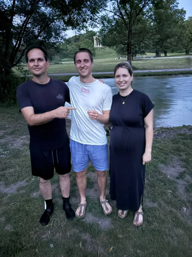 Drei Personen am Schwabinger Bach im Englischen Garten, im Hintergrund der Monopteros, ein Mann hält den gefundenen Ring hoch