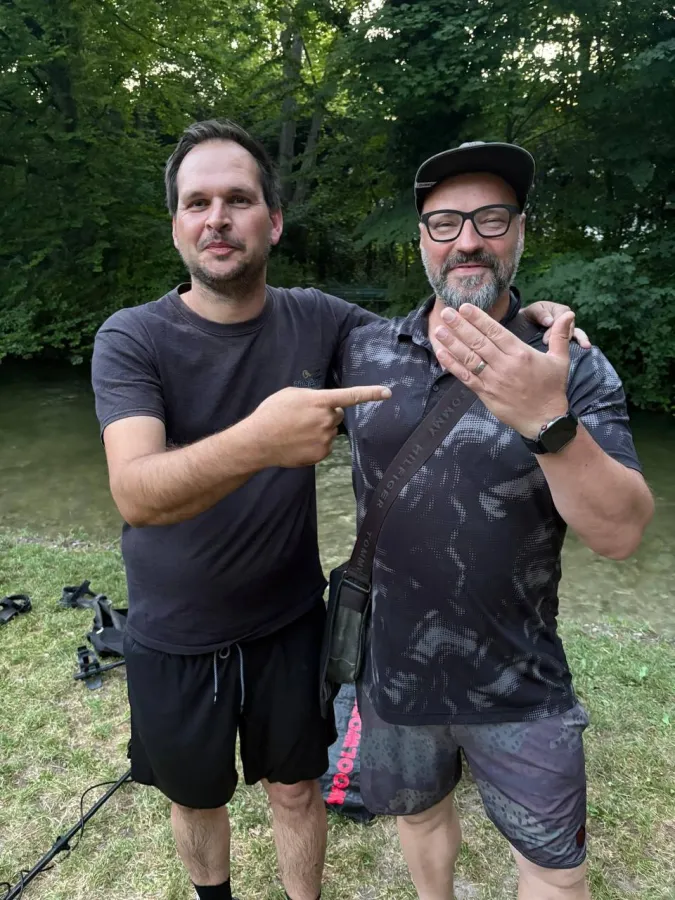 Zwei Männer am Ufer des Schwabinger Bachs im Englischen Garten, einer zeigt auf den wiedergefundenen Ehering am Finger des anderen